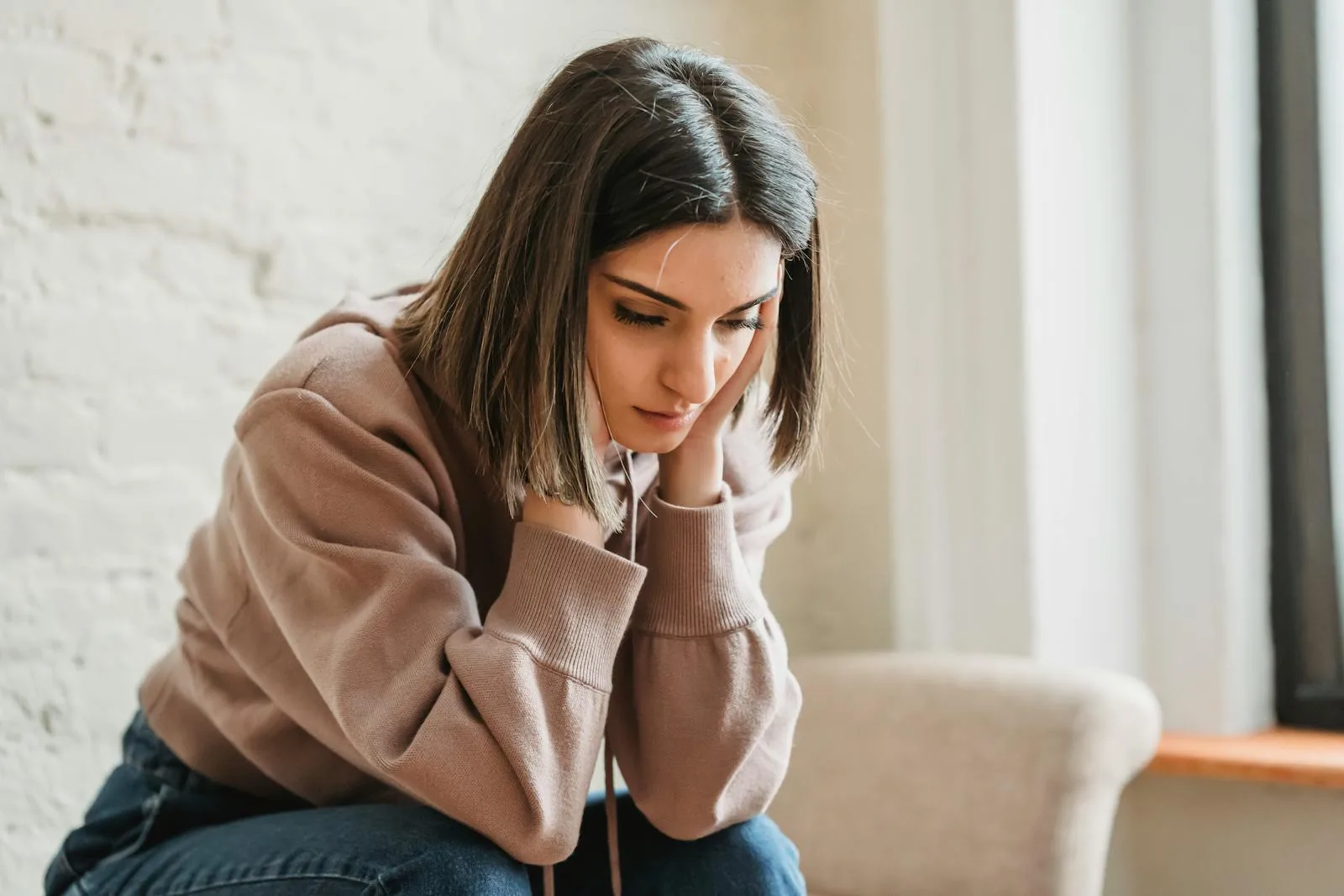 Unhappy female in casual wear touching face and looking down while sitting on sofa in light living room at home.