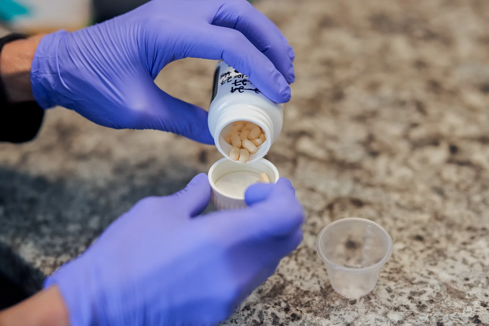 Gloved clinician pouring medication tablets from a labeled bottle into a dosing cup.