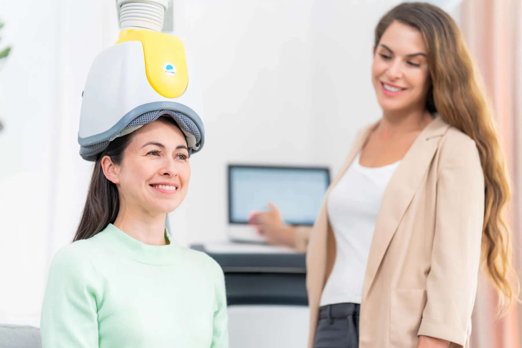 A smiling patient wearing the BrainsWay yellow H7 Deep TMS helmet while a trained AriseVida Care clinician guides the session in the background.